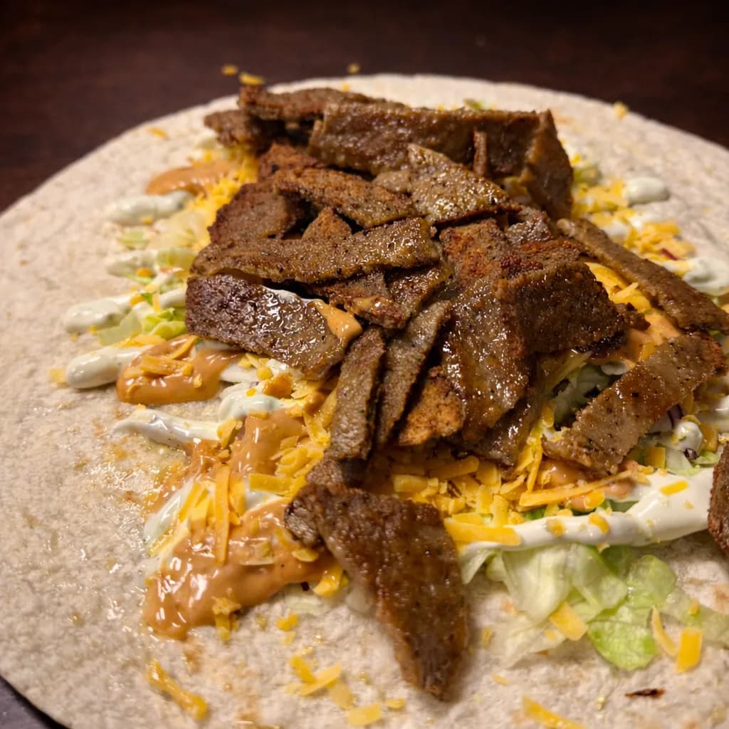 Doner kebab wrap being prepared with sliced doner meat, shredded lettuce, grated cheese, and drizzled with garlic and chili sauce on a flatbread.